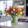 British tulips in a glass vase placed on a kitchen island. The tulips are in mixed colours like yellow, pink, purple, white and red.
