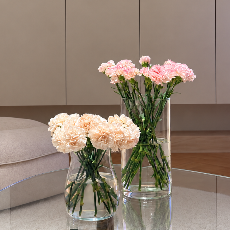 Two clear glass vases filled with pink and cream carnation flowers arranged on a round glass-top coffee table in a modern living room with beige cabinetry and wood flooring.