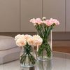 Two clear glass vases filled with pink and cream carnation flowers arranged on a round glass-top coffee table in a modern living room with beige cabinetry and wood flooring.