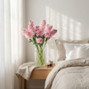 Pink flowers arranged in a clear glass vase on a wooden bedside table next to a bed with beige linens and white pillows, softly lit by window shadows.
